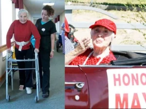 An elderly woman, using a walker, assisted by a younger woman in medical scrubs in a hallway. 